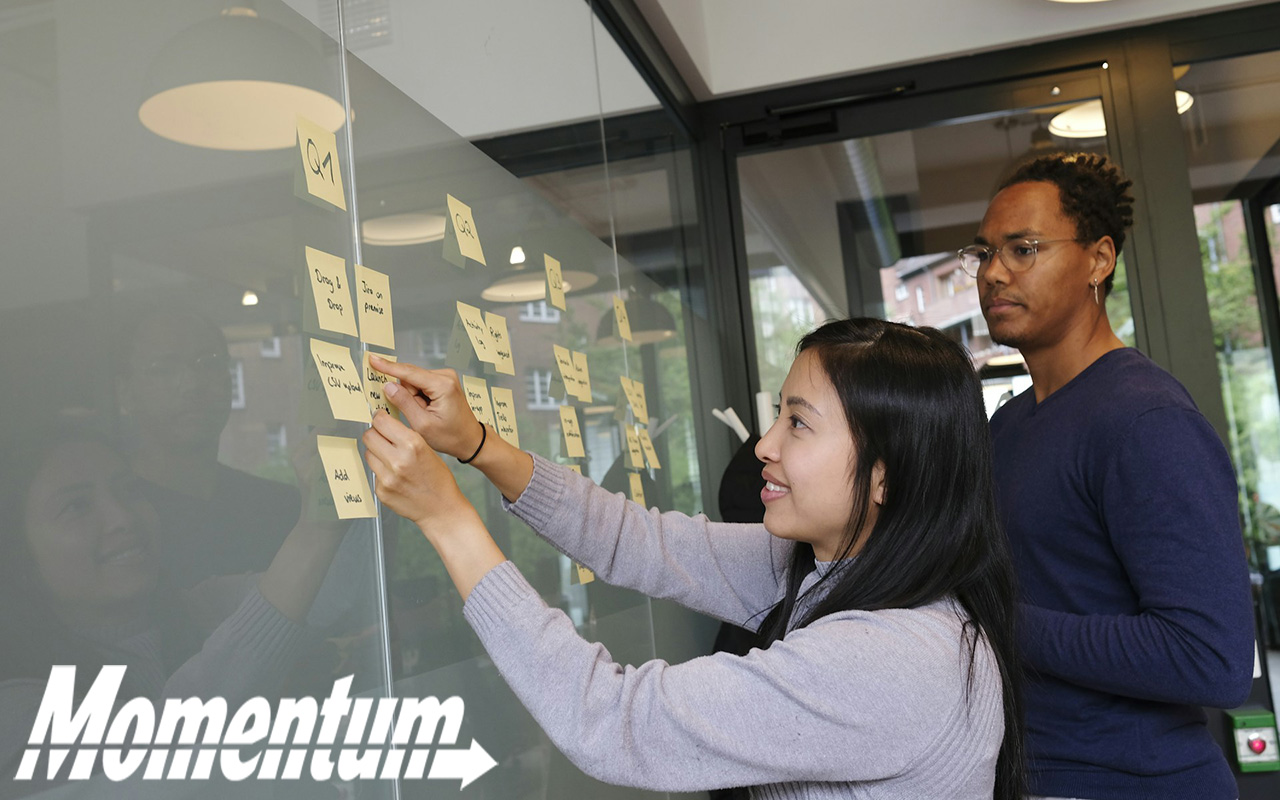 An Asian woman and a black man add sticky notes to a presentation board.