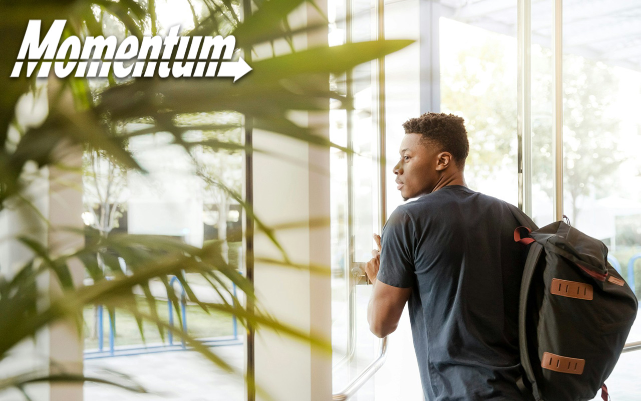 Black male student, wearing a backpack, exiting a building through a glass set of doors. 