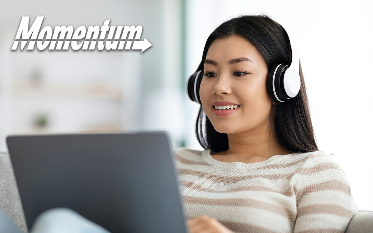 Young Asian woman wearing headphones while she looks at a laptop propped up in her knees.